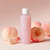 A pink bottle of Anua Peach 77% Niacin Essence Toner with peach fruit extracts, alongside fresh peaches on a pink background.