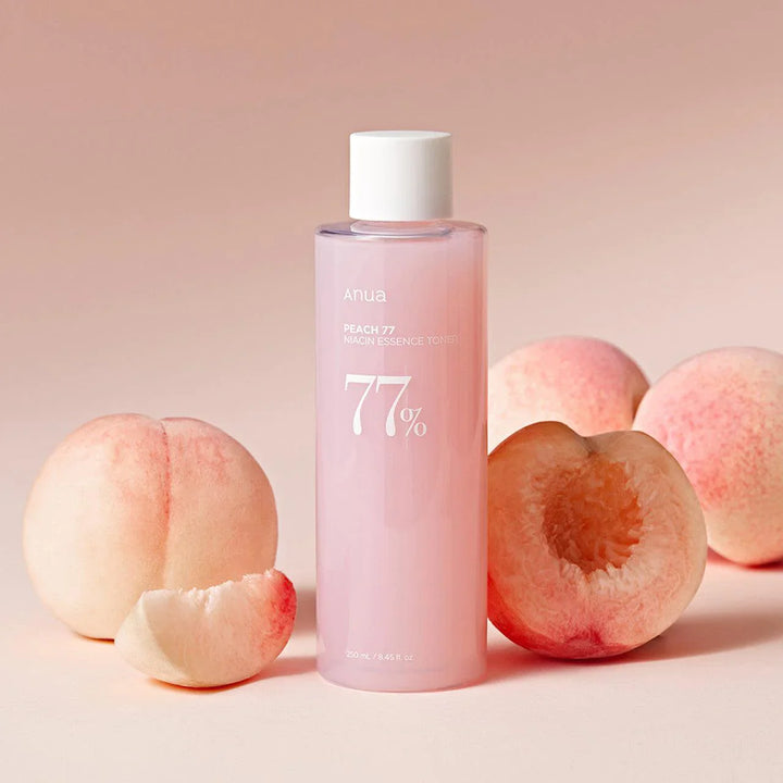 A pink bottle of Anua Peach 77% Niacin Essence Toner with peach fruit extracts, alongside fresh peaches on a pink background.